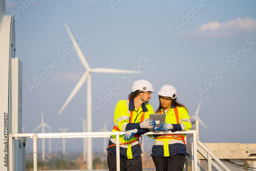 Caucasian engineer or technician man and woman discuss together and they stay on base of windmill in front of row of wind turbine with blue sky and warm light.