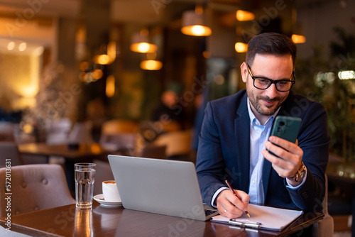 Happy smiling businessman wearing suit using laptop and smartphone about to make a deal while sitting and remotely working in modern city restaurant writing notes on clipboard satisfied with the offer