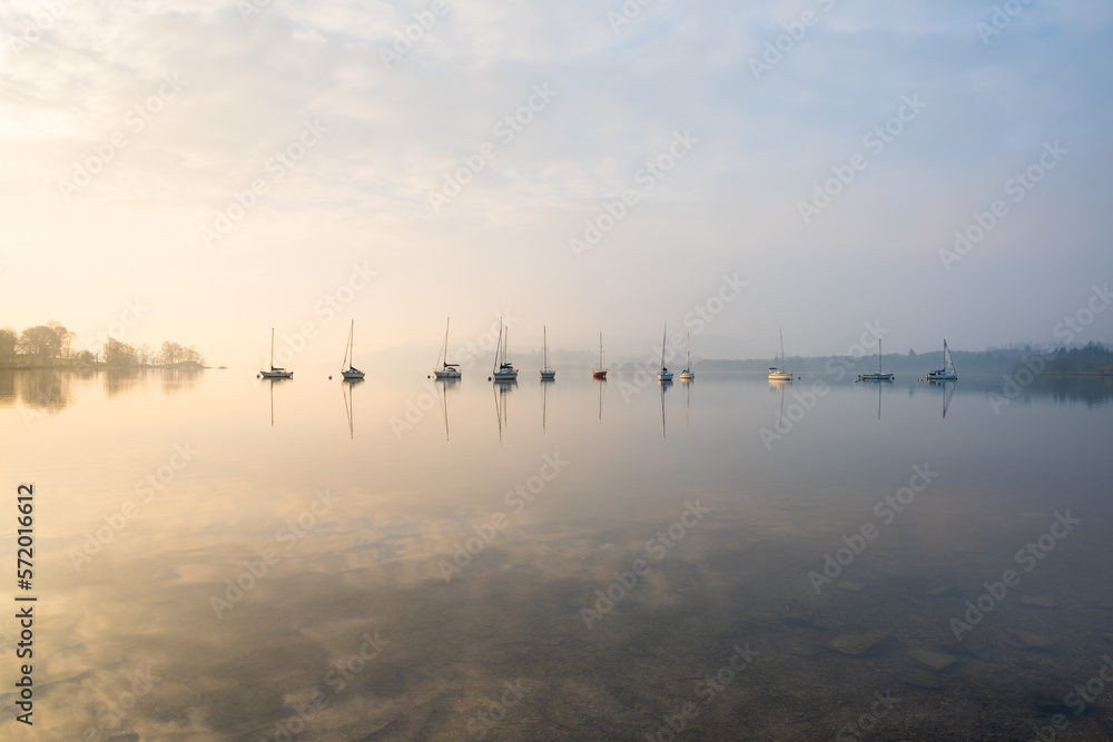 Row of yachts sailing boats on a misty morning at Windermere in The Lake District, UK with calm reflections in the water. Row of yachts sailing boats on a misty morning at Windermere in The Lake District, UK with calm reflections in the water.