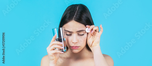 Brunette take some pills, holds glass of water, isolated on blue. Brunette taking a pill with a glass of water. Woman taking drugs to releave headache photo