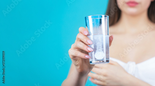 Woman taking drugs to releave headache. Brunette take some pills, holds glass of water, isolated on blue photo