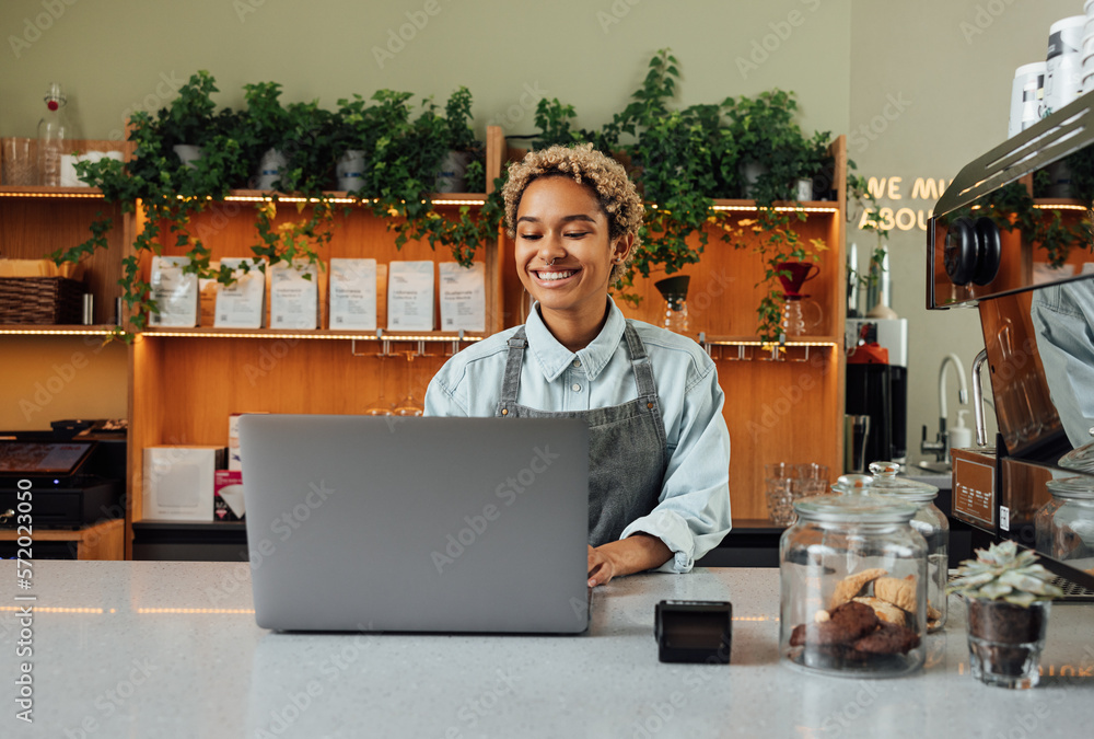 Smiling barista in apron typing on laptop at the counter. Cheerful coffee shop owner working while standing at the bar counter. Smiling barista in apron typing on laptop at the counter. Cheerful coffee shop owner working while standing at the bar counter.