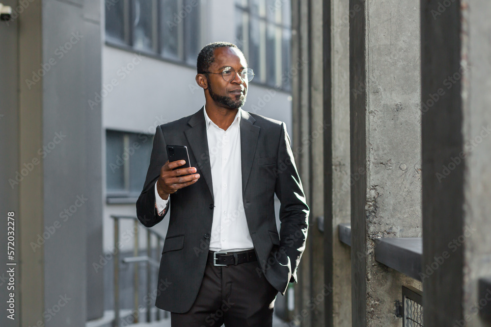 African American businessman in suit standing outside near office, bank, holding phone, hand in pocket. He is waiting for a business meeting, a client, a partner. African American businessman in suit standing outside near office, bank, holding phone, hand in pocket. He is waiting for a business meeting, a client, a partner.