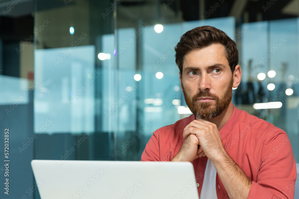 Close-up photo. A young male programmer, designer, freelancer in a red shirt who sits and works in the office at a laptop. He folded his hands together, looks thoughtfully and seriously to the side. Close-up photo. A young male programmer, designer, freelancer in a red shirt who sits and works in the office at a laptop. He folded his hands together, looks thoughtfully and seriously to the side.