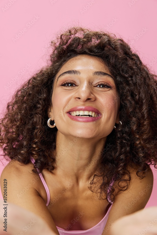 Woman with curly afro hair dancing in pink top and jeans on a pink background, smiling, stretching her arms into the camera, copy space