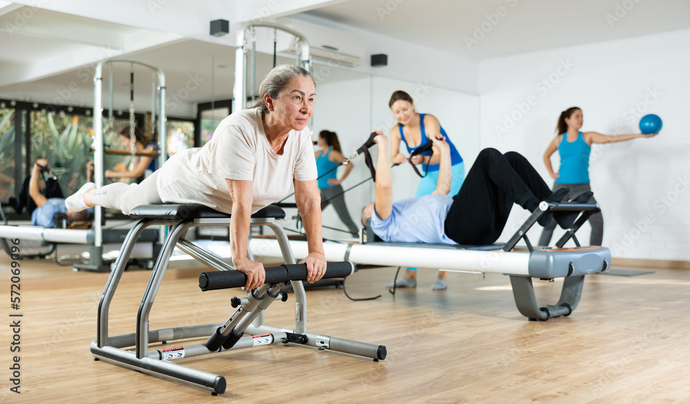 Willing aged woman engaging in pilates training on pedal fitness chair in exercise room during workout session. Persons practicing pilates with trainer