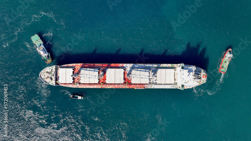 Aerial drone top down photo of super tanker towed by tug boat in deep blue sea near logistics container terminal port