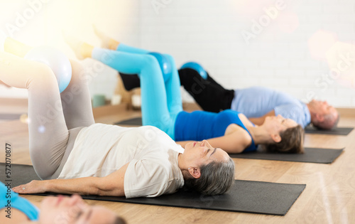 Diligent elderly woman practicing pilates with ball in exercise room during pilates classes. Persons doing pilates in fitness hall.