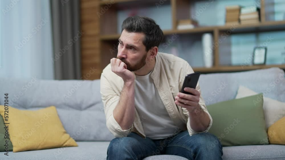 Boring tired bearded man scrolling feed of social networks products in internet store on smartphone sitting on couch in living room Exhausted sad male look at mobile screen without enthusiasm indoor