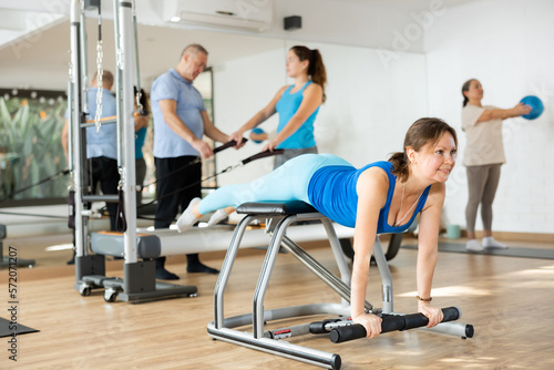 Slender middle-aged woman practicing pilates on pedal fitness chair in sports hall during pilates classes. Persons doing pilates with trainer