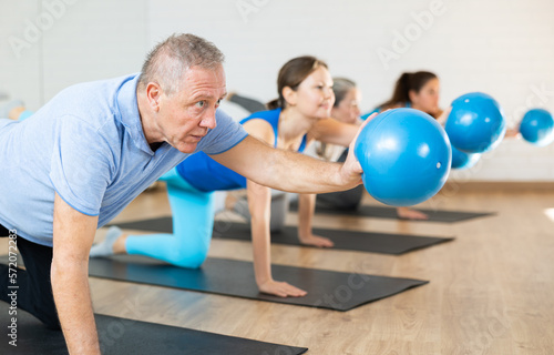 Aged man maintaining active lifestyle exercising with small pilates ball during group class in modern fitness center
