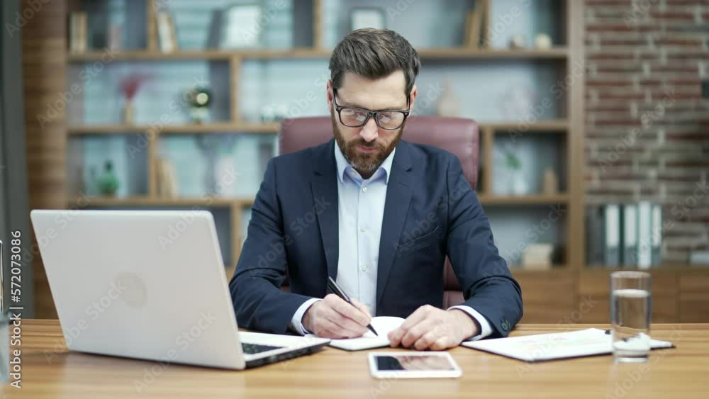 Busy mature business man investor with glasses write notes at organizer planning workday to do list or meeting schedule and typing on laptop computer working online at desktop in modern office indoors Busy mature business man investor with glasses write notes at organizer planning workday to do list or meeting schedule and typing on laptop computer working online at desktop in modern office indoors