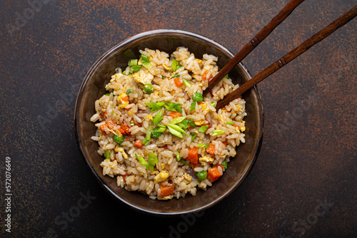 Authentic Chinese and Asian fried rice with egg and vegetables in ceramic brown bowl top view on dark rustic concrete table background. Traditional dish of China