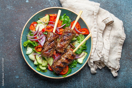 Grilled skewer meat beef kebabs on sticks served with fresh vegetables salad on plate on rustic concrete background from above. Traditional Middle Eastern and Turkish dish Kebab