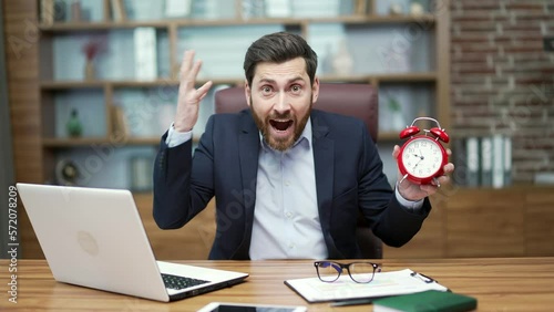 Nervous displeased bearded mature business man looking at camera holding in hands and pointing at clock asking to hurry up and warning about deadline sitting next to laptop computer at modern office photo