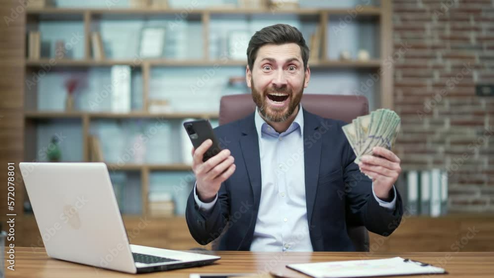 Overjoyed happy mature business man investor receive message with great news at desk in office Successful entrepreneur holding money and rejoices at good earnings income salary investment at workplace Overjoyed happy mature business man investor receive message with great news at desk in office Successful entrepreneur holding money and rejoices at good earnings income salary investment at workplace