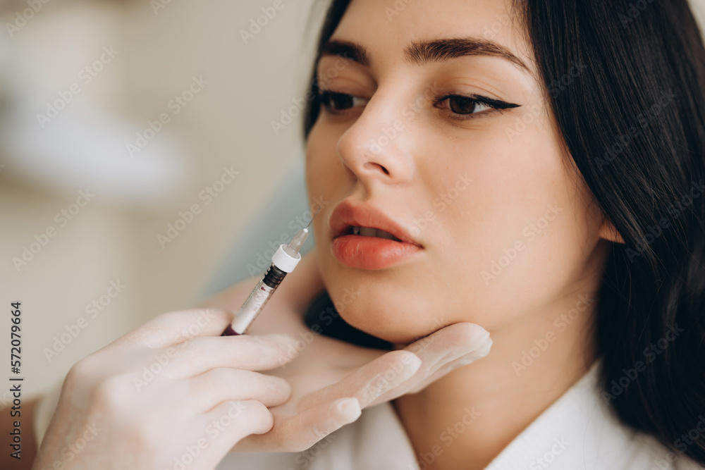 Close up of hands of cosmetologist making botox injection in female lips. She is holding syringe. The young beautiful woman is receiving procedure with enjoyment. Close up of hands of cosmetologist making botox injection in female lips. She is holding syringe. The young beautiful woman is receiving procedure with enjoyment.