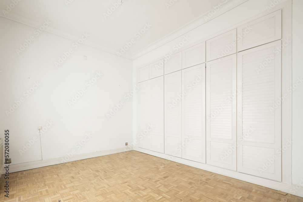 Empty room with one wall covered with white wood cabinets, mezzanine chests all with white Venetian type doors, plain white painted walls and checkerboard shaped oak slatted floors Empty room with one wall covered with white wood cabinets, mezzanine chests all with white Venetian type doors, plain white painted walls and checkerboard shaped oak slatted floors