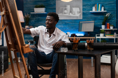 Smiling african american male cartoonist using colored pencils to sketch on easel at art studio. Wheelchair user drawing happily on canvas in bedroom. Person with artistic skills working in atelier.