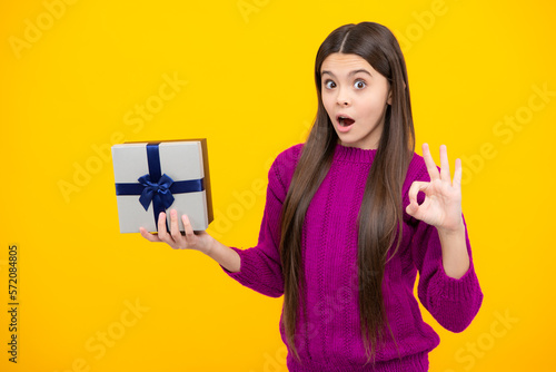 Surprised teenage child holding gift box on yellow isolated background. Gift for kids birthday. Christmas or New Year present box. Surprised emotions of young teenager girl.