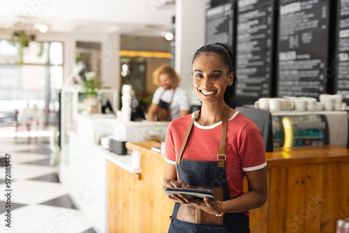 Portrait of happy biracial female barista wearing apron and using tablet in cafe photo