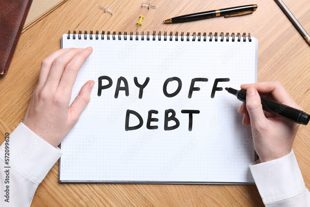 Woman writing phrase Pay Off Debt in notebook at wooden table, top view Woman writing phrase Pay Off Debt in notebook at wooden table, top view