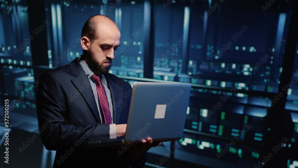 IT server engineer inspecting working computer processor with laptop, standing in professional data center. Infrastructure specialist running hosting server diagnostics. Tripod shot. IT server engineer inspecting working computer processor with laptop, standing in professional data center. Infrastructure specialist running hosting server diagnostics. Tripod shot.