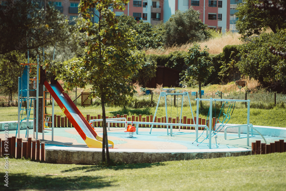 The capture of a children's playground, focusing on a colorful slide next to the swing and the climbing equipment in a round area partially enclosed by wood stakes The capture of a children's playground, focusing on a colorful slide next to the swing and the climbing equipment in a round area partially enclosed by wood stakes