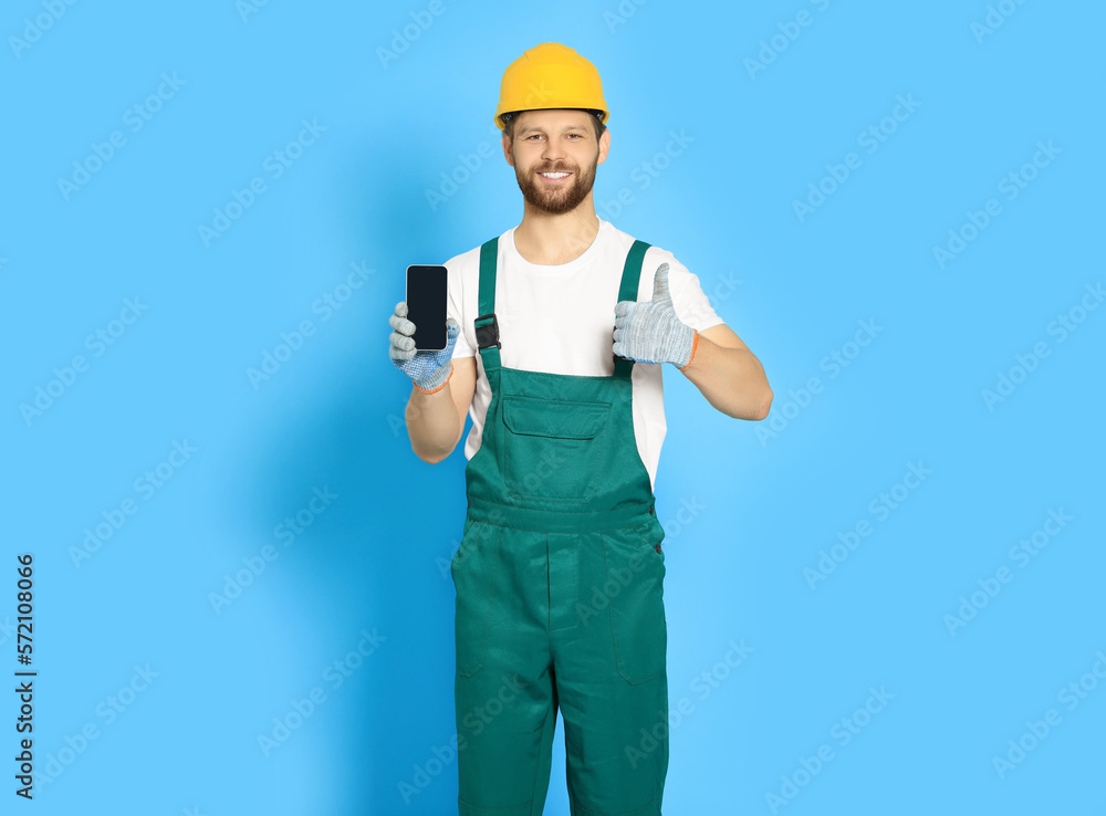 Professional repairman in uniform showing smartphone and thumbs up on light blue background Professional repairman in uniform showing smartphone and thumbs up on light blue background