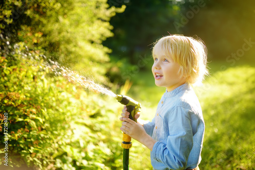 Funny little boy watering plants and playing with garden hose with sprinkler in sunny backyard. Preschooler child having fun with spray of water. Summer outdoors activity for kids