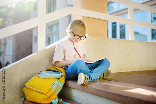 Little student doing homework on break on stair of elementary school building. Portrait of funny nerd schoolboy with big glasses. Vision problems. Back to school concept. Education for kids.