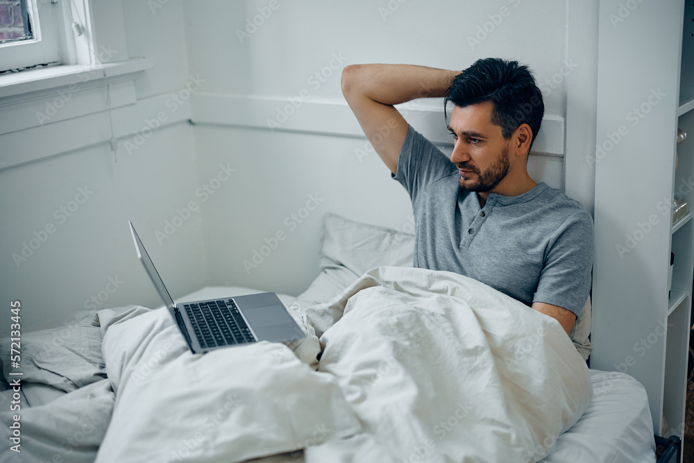 Obraz premium Mid adult man using laptop while relaxing in bedroom.