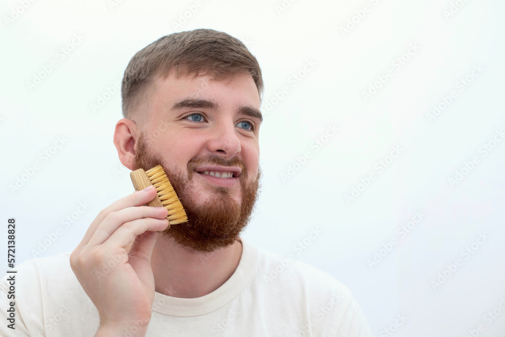 Handsome young bearded guy, European brutal man with a beard hair is combing the beard on face with a comb, brush. Male personal self care, products, accessory isolated on white background Handsome young bearded guy, European brutal man with a beard hair is combing the beard on face with a comb, brush. Male personal self care, products, accessory isolated on white background