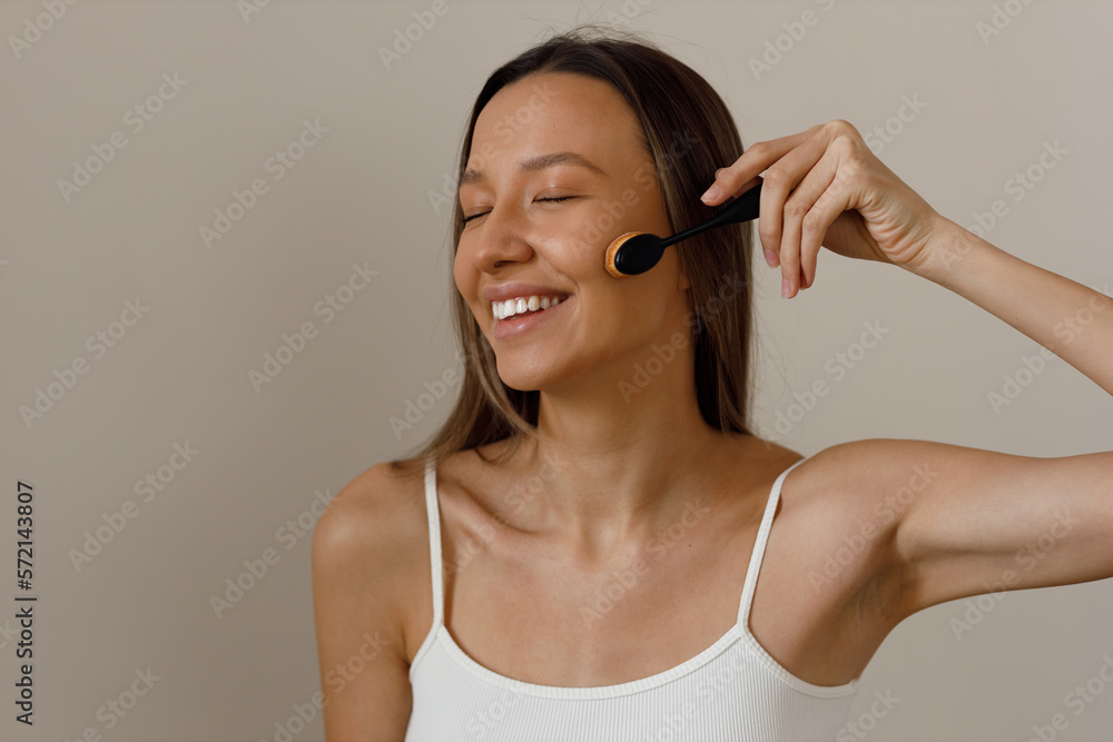 Beautiful young smiling blonde woman with long hair, dressed in short white top, apply foundation on face. Applying light layer of matting powder using professional brush. Beautiful young smiling blonde woman with long hair, dressed in short white top, apply foundation on face. Applying light layer of matting powder using professional brush.