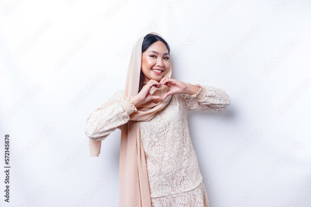 A happy young Asian woman wearing hijab feels romantic shapes heart gesture expresses tender feelings