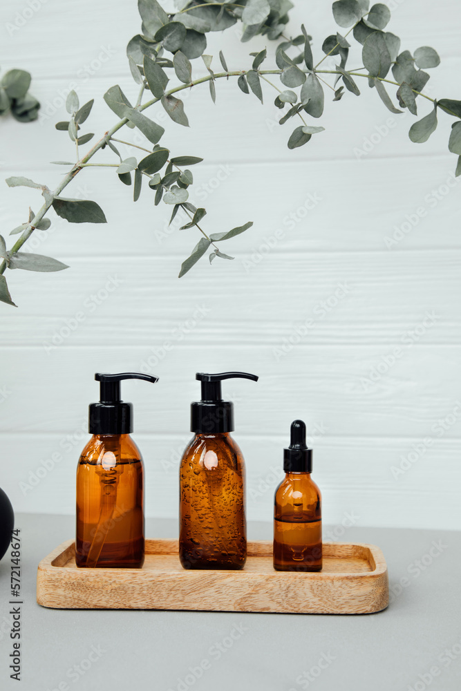 Amber bottles with facial cosmetics on a tray with a branch of eucalyptus on the gray wooden background, facial cleaner, serum. Front view Amber bottles with facial cosmetics on a tray with a branch of eucalyptus on the gray wooden background, facial cleaner, serum. Front view