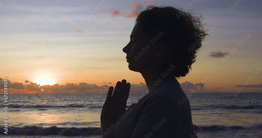 Silhouette of woman at sunset, next to sea waves and blown by warm wind. Calm Caucasian woman mentally reading prayer to God. Religious woman stands on seashore, lays hands in prayer. Concept of hope. Silhouette of woman at sunset, next to sea waves and blown by warm wind. Calm Caucasian woman mentally reading prayer to God. Religious woman stands on seashore, lays hands in prayer. Concept of hope.