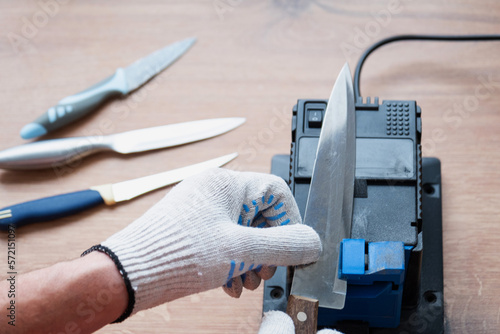 Sharpening a knife on an electric sharpener at home. The man's hand drives the knife blade between the blue sharpeners, dust flies on the machine.