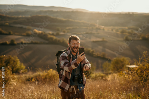 A young hiker man standing on the mountain peak, having a video call and showing the landscape. Golden hour