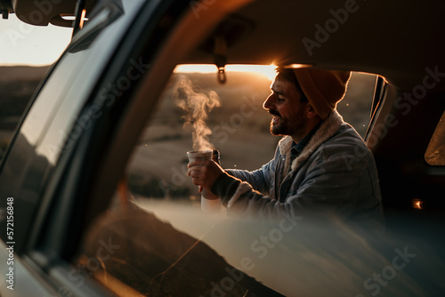 A smiling man sits in the trunk of a car and drinks a fresh hot coffee by himself.