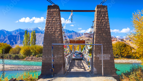 Suspension bridge over green Indus river.The way to Stakna gompa with fall foliage in Leh, Ladakh, India. Narrow bridge with car. Green river and winter blue sky. Dangerous highway in Karakorum range. photo