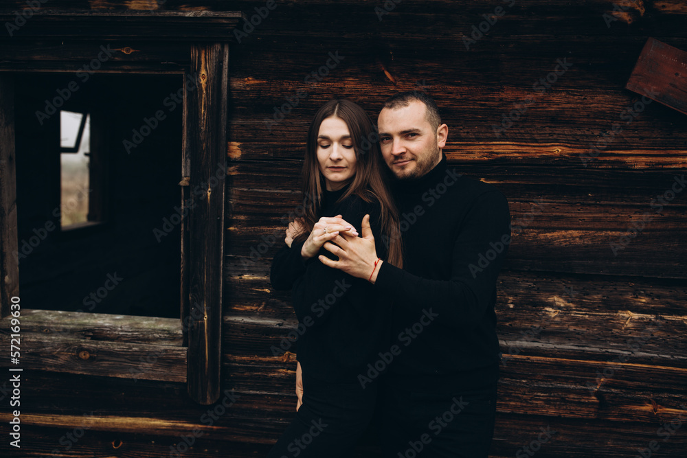 a man and a woman in black clothes near an old wooden house. photo in dark brown tones. old abandoned house and cloudy cold weather. beautiful stylish couple on the background of a brown wooden wall. a man and a woman in black clothes near an old wooden house. photo in dark brown tones. old abandoned house and cloudy cold weather. beautiful stylish couple on the background of a brown wooden wall.