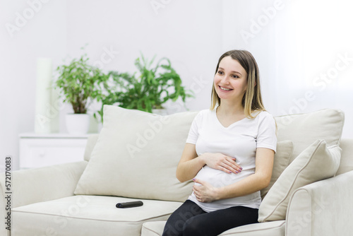 Pregnant woman sitting on white sofa and touching her stomach.