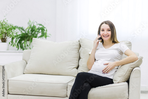 Smiling pregnant woman talking over the phone on white sofa.