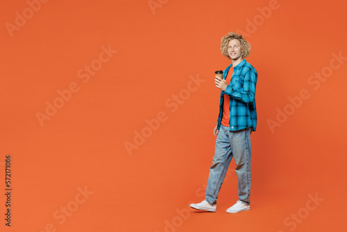 Full body side view young blond man wear blue shirt orange t-shirt hold takeaway delivery craft paper brown cup coffee to go isolated on plain red background studio portrait. People lifestyle concept.