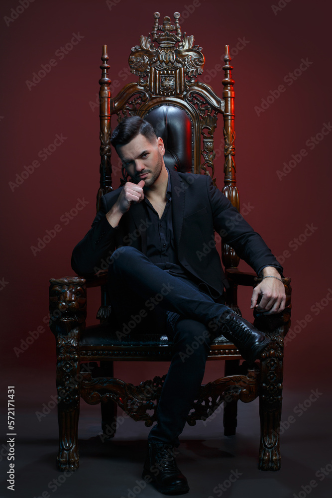 Elegant man in a classic  suit sitting in a carved wooden chair against a dark background.