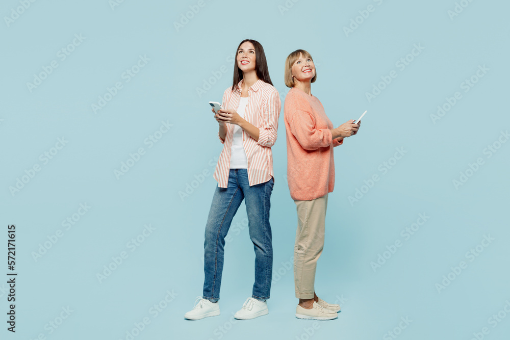 Full body fun happy elder parent mom with young adult daughter two women together wear casual clothes hold in hand mobile cell phone look overhead isolated on plain blue background Family day concept