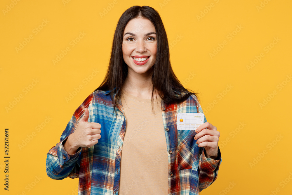 Young cheerful fun smiling woman wear blue shirt beige t-shirt holding in hand mock up of credit bank card show thumb up isolated on plain yellow background studio portrait. People lifestyle concept. Young cheerful fun smiling woman wear blue shirt beige t-shirt holding in hand mock up of credit bank card show thumb up isolated on plain yellow background studio portrait. People lifestyle concept.