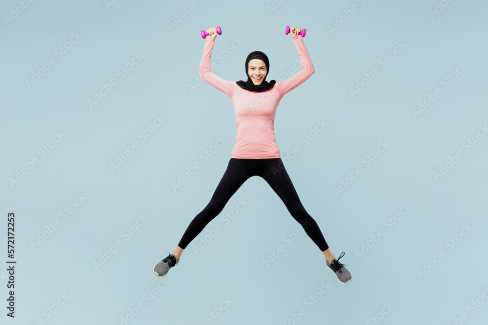 Full body young asian muslim fitness trainer sporty woman wear pink abaya hijab spend time in home gym jump high hold dumbbells isolated on plain blue background studio. Workout sport fit abs concept. Full body young asian muslim fitness trainer sporty woman wear pink abaya hijab spend time in home gym jump high hold dumbbells isolated on plain blue background studio. Workout sport fit abs concept.