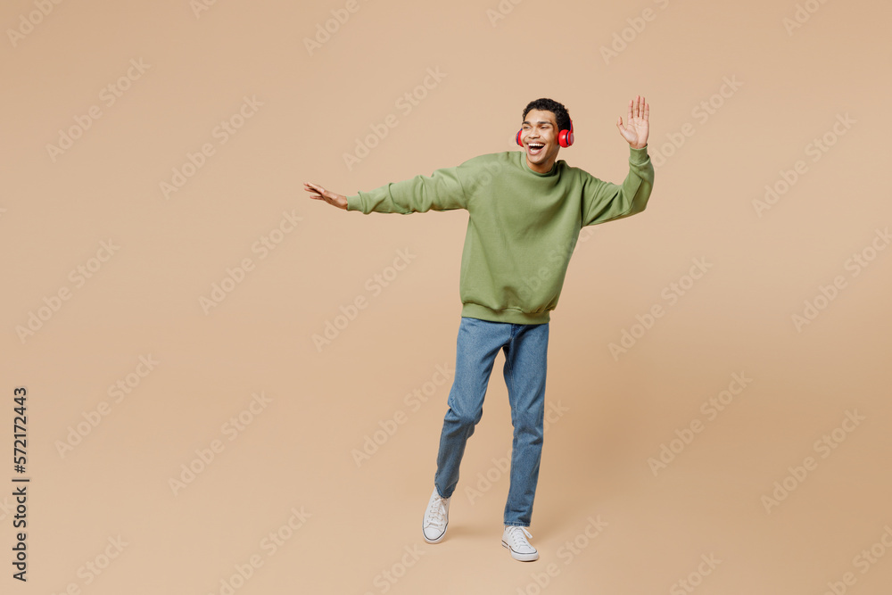 Full body cheerful fun young man of African American ethnicity wear green sweatshirt headphones listen to music dancing raise up hands isolated on plain pastel light beige background studio portrait. Full body cheerful fun young man of African American ethnicity wear green sweatshirt headphones listen to music dancing raise up hands isolated on plain pastel light beige background studio portrait.